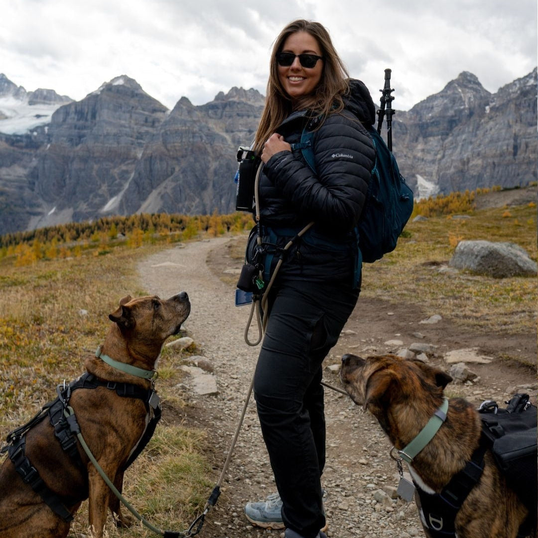 Woman wearing an adjustable hands-free leash over her shoulder, walking two dogs with a scenic mountain landscape in the background.